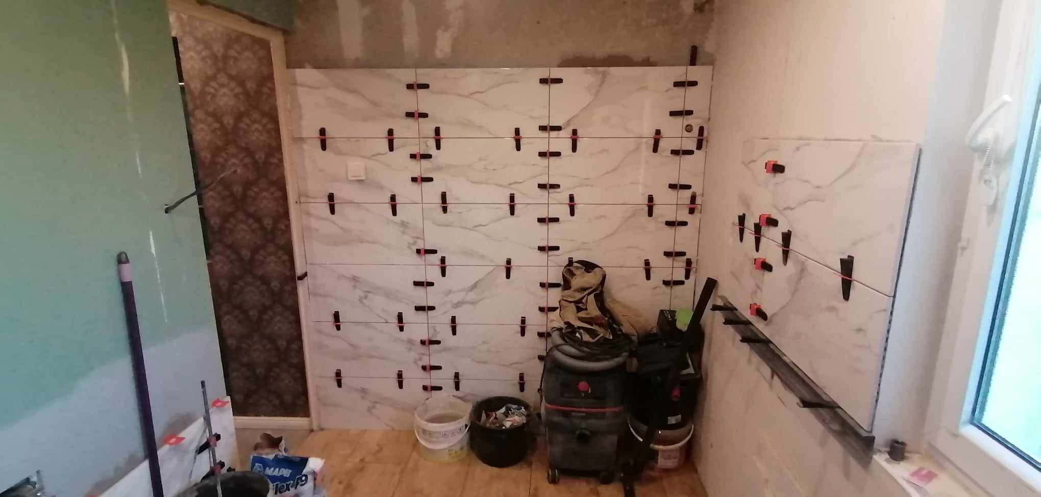 Układanie płytek imitujących marmur na ścianie w łazience, z użyciem systemu poziomowania. Widoczne narzędzia i akcesoria glazurnicze.