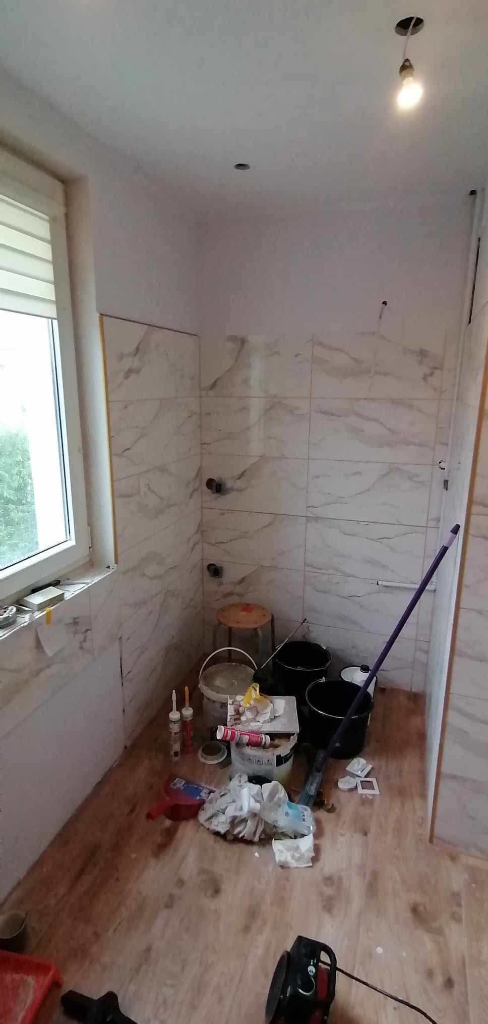 Wnętrze łazienki w trakcie wykańczania: białe płytki imitujące marmur na ścianach, widoczne narzędzia i materiały budowlane, okno z żaluzjami. Częściowo ułożona glazura.
