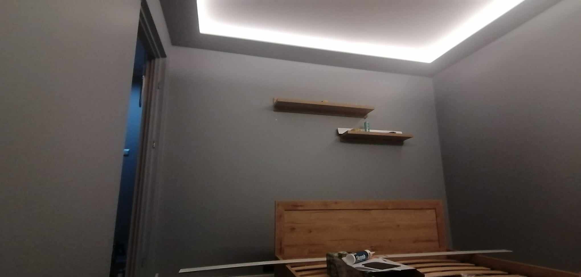 Szara sypialnia po remoncie: podświetlany sufit, drewniane półki i rama łóżka. Widoczny fragment otwartych drzwi. Świeżo pomalowane ściany.