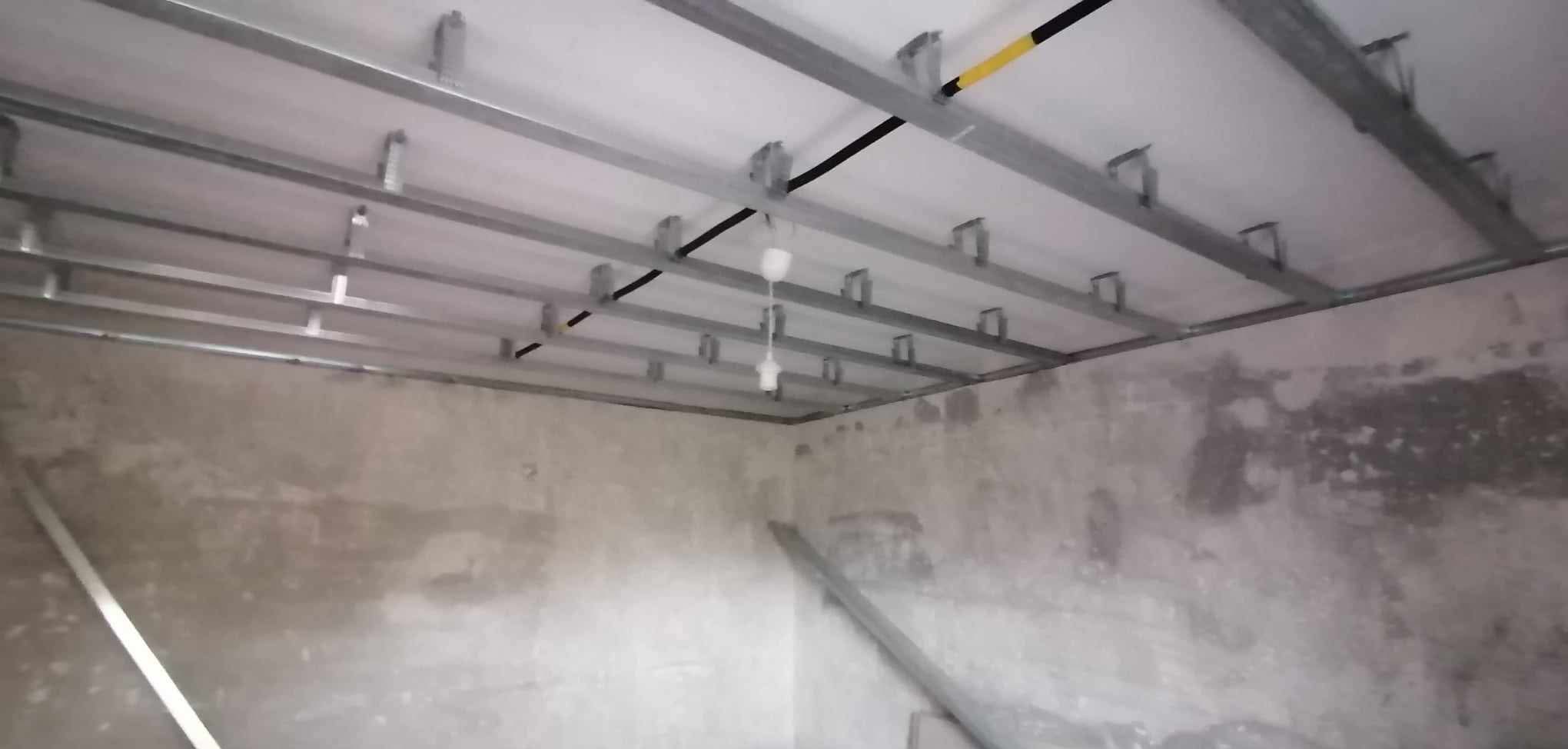 Metalowa konstrukcja sufitu podwieszanego z płyt karton-gips, widok z dołu na szkielet przed montażem płyt, surowe ściany w tle.