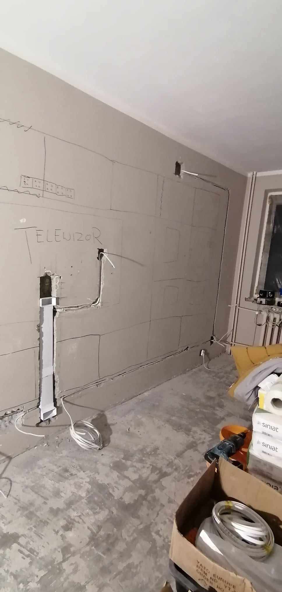 Ściana z płyt karton-gips z wyrysowanym miejscem na telewizor i poprowadzonymi kablami. Widoczne narzędzia i materiały budowlane. Etap prac wykończeniowych.