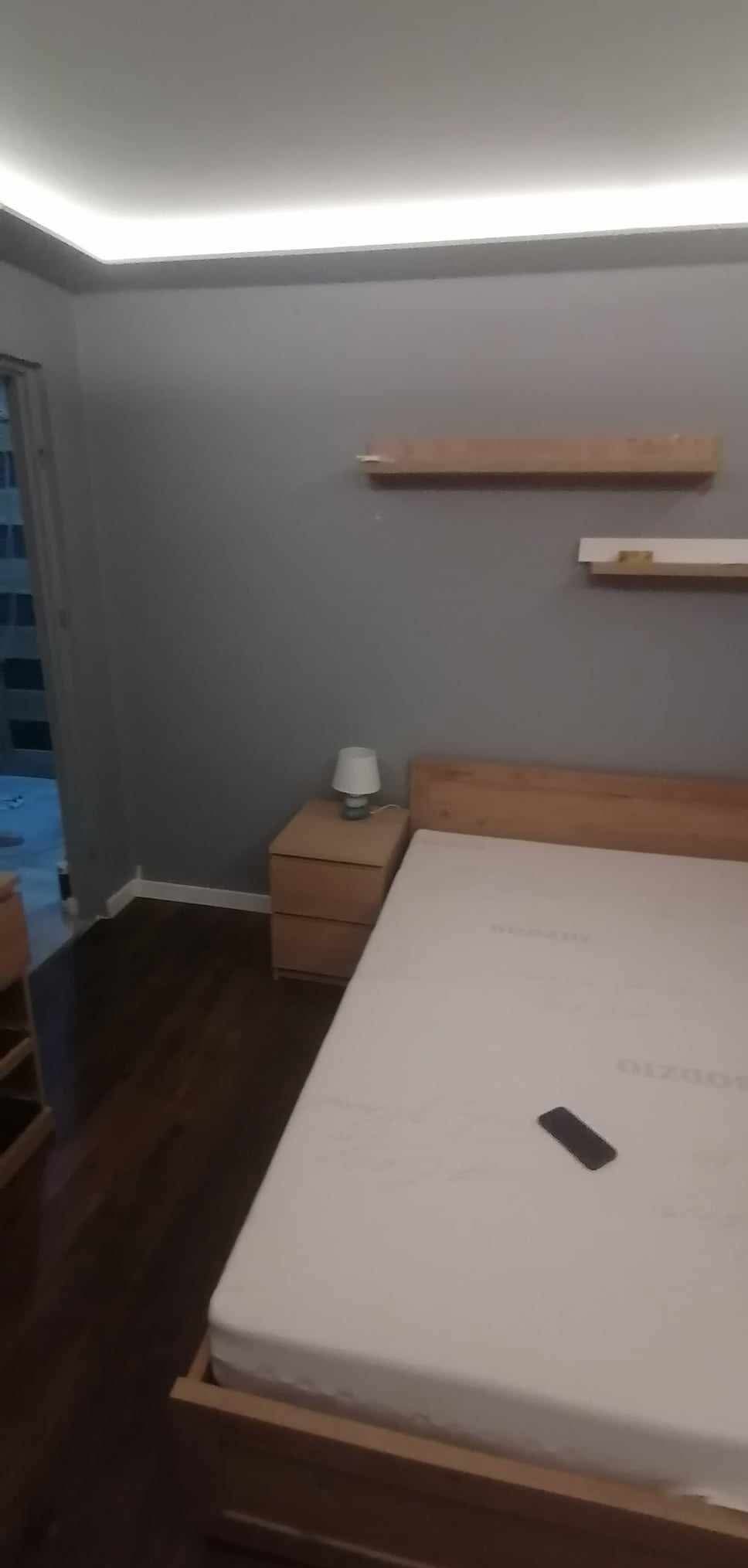 Sypialnia po remoncie: drewniane półki na szarej ścianie, podświetlany sufit, drewniane łóżko z białą pościelą i stolik nocny z lampką. Ciemna podłoga.