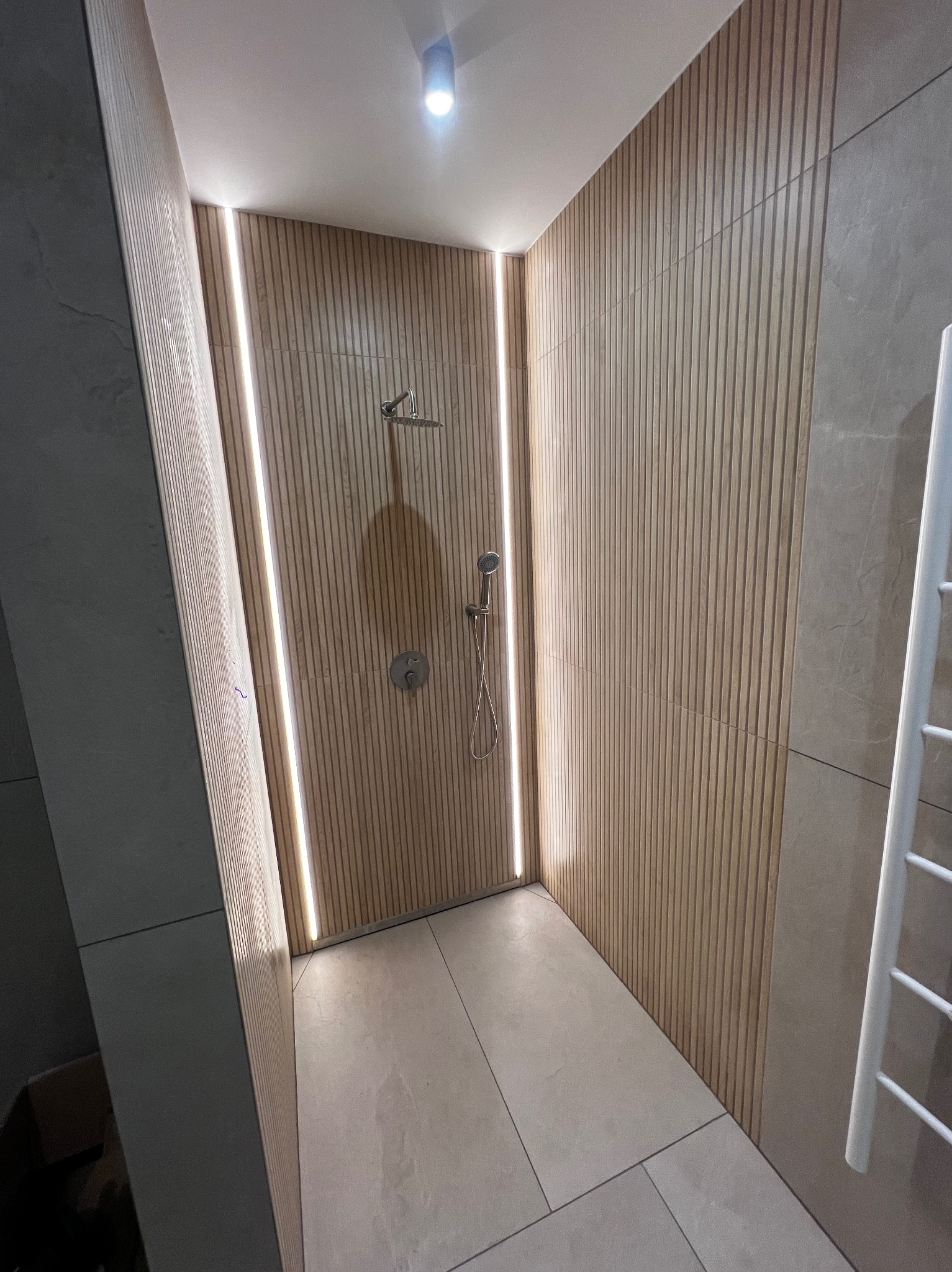 Nowoczesna kabina prysznicowa z drewnopodobnymi panelami ściennymi i subtelnym oświetleniem LED. Minimalistyczny design, beżowa kolorystyka, widoczny fragment grzejnika.