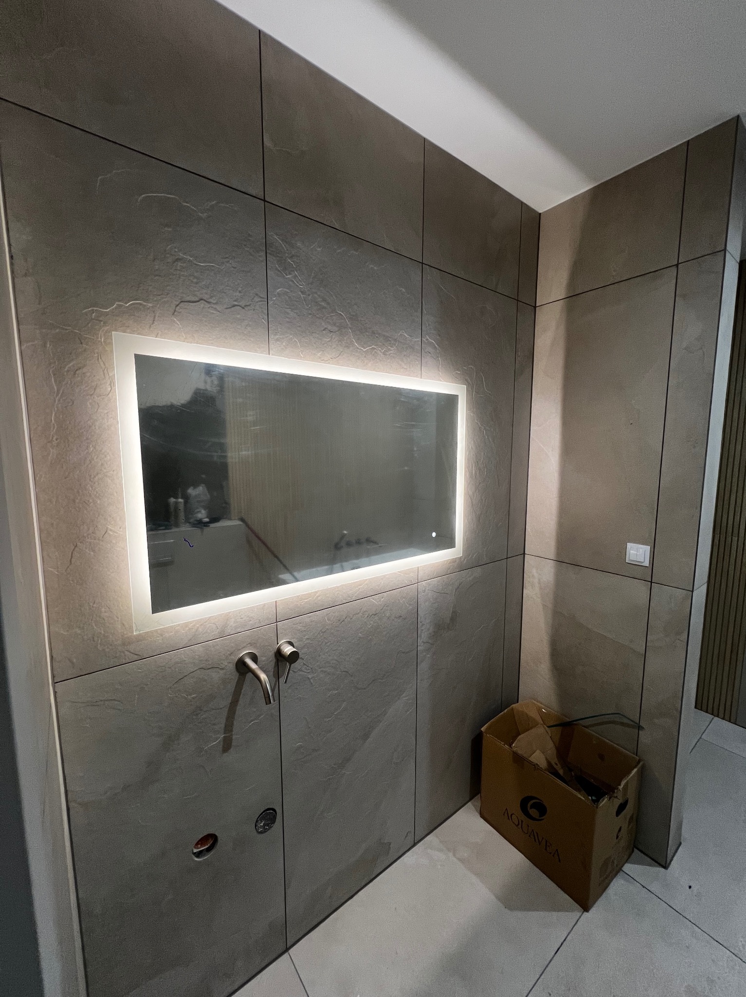 Wykończona łazienka z szarymi płytkami imitującymi kamień, podświetlanym lustrem i minimalistyczną armaturą. Widoczny karton po produkcie.