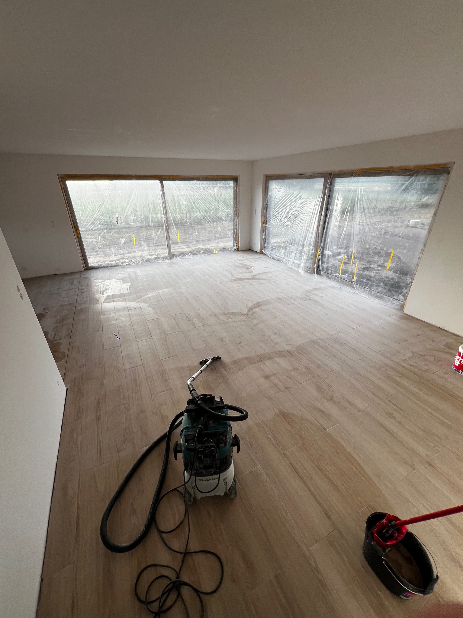 Jasne płytki podłogowe imitujące drewno w przestronnym pokoju z dużymi oknami zabezpieczonymi folią. Widoczny odkurzacz przemysłowy i wiadro z mieszadłem.