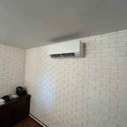 Grzegorz Ocias - Biały klimatyzator na ścianie z cegły w jasnym wnętrzu. Widoczny fragment komody. Ujęcie z dołu, podkreślające montaż urządzenia.