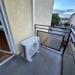 Grzegorz Ocias - Biała jednostka zewnętrzna klimatyzacji zamontowana na szarym balkonie z widokiem na budynki i niebo. Widoczne elementy mocujące i fragment ściany budynku.