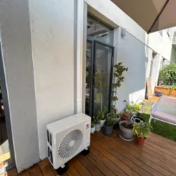 Biały klimatyzator zewnętrzny marki Gree zamontowany na drewnianym tarasie przy ścianie budynku, obok okna i donic z roślinami.