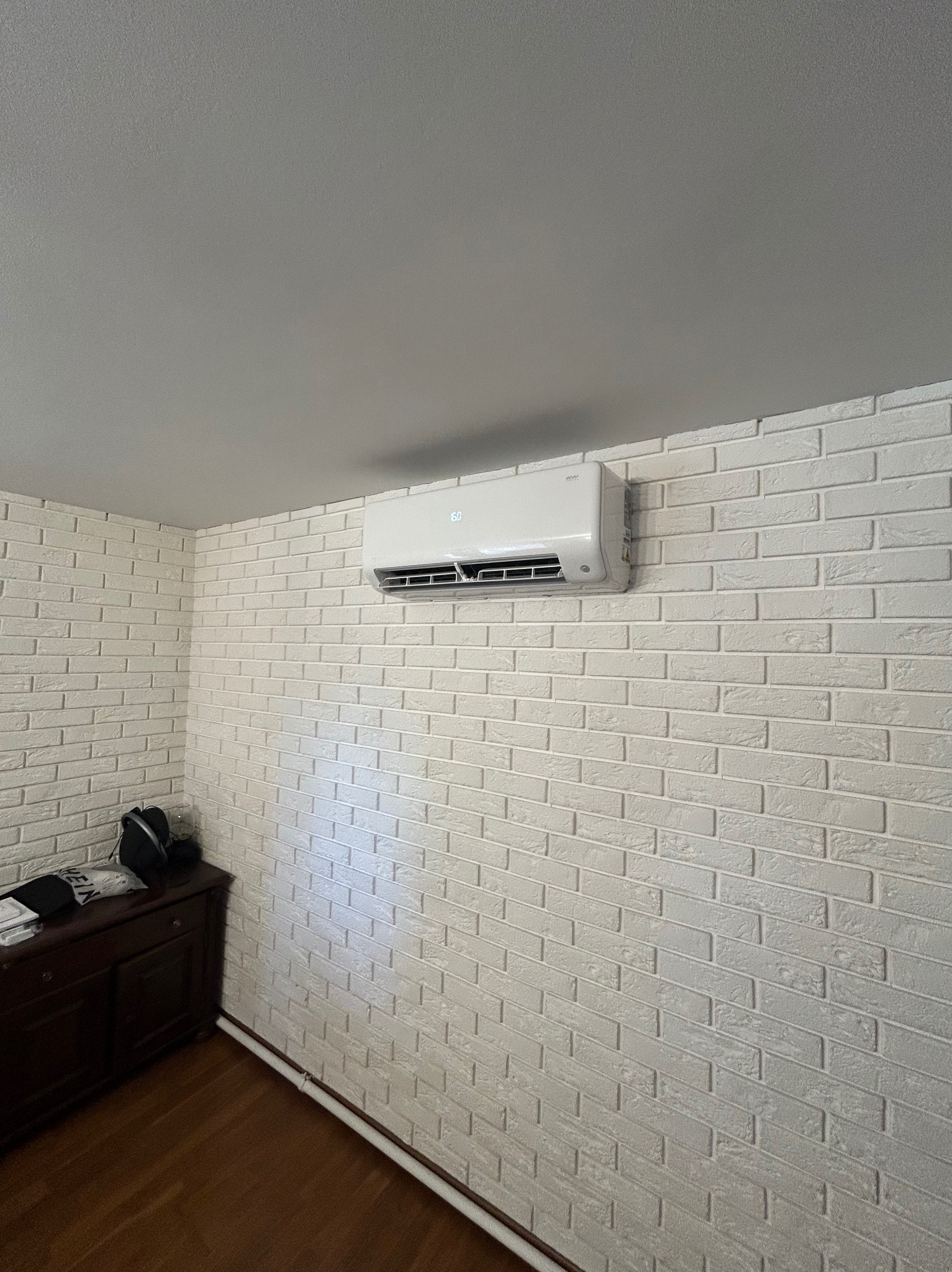 Biały klimatyzator na ścianie z cegły w jasnym wnętrzu. Widoczny fragment komody. Ujęcie z dołu, podkreślające montaż urządzenia.