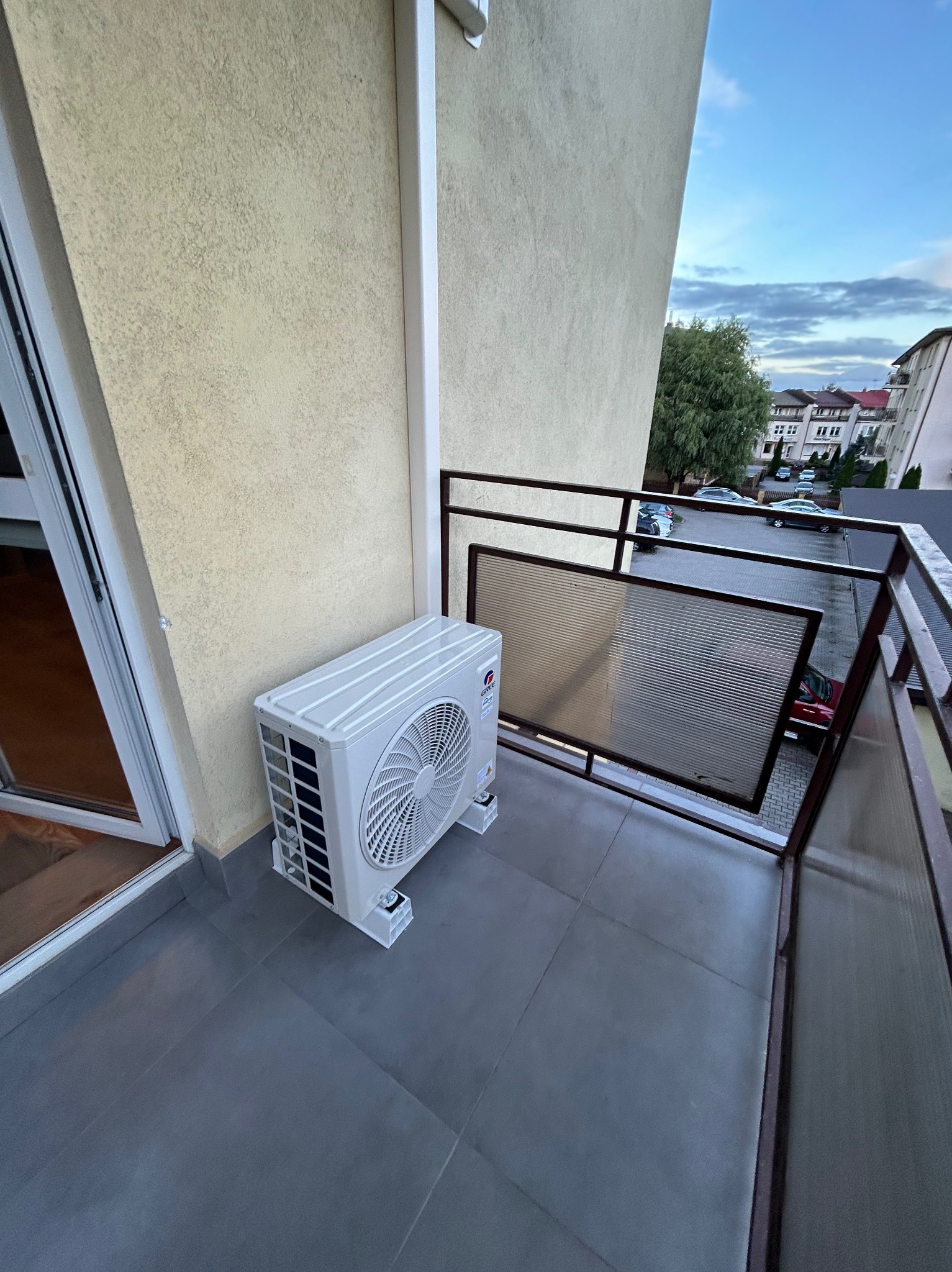 Biała jednostka zewnętrzna klimatyzacji zamontowana na szarym balkonie z widokiem na budynki i niebo. Widoczne elementy mocujące i fragment ściany budynku.