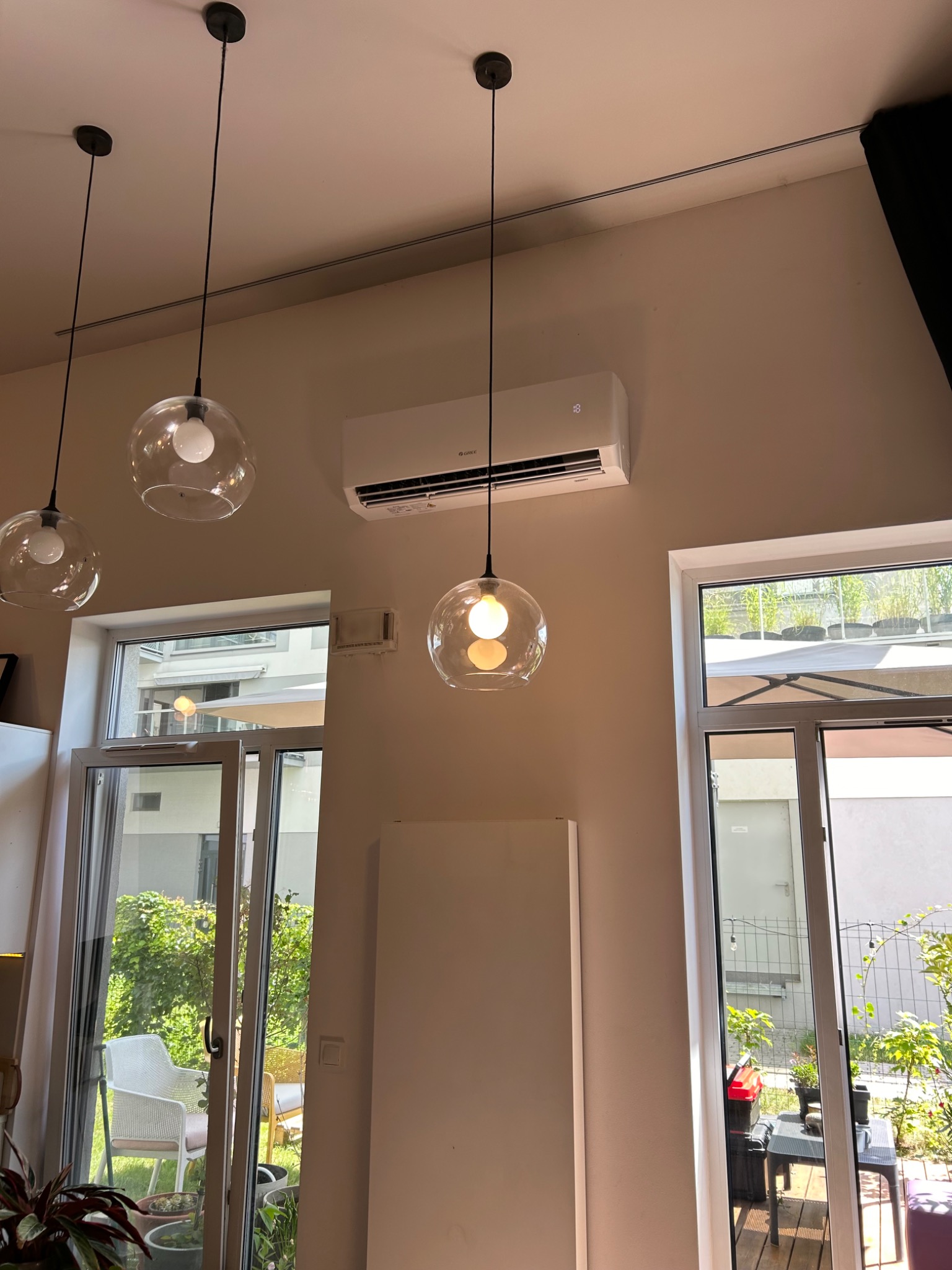 Biała klimatyzacja ścienna Gree w nowoczesnym wnętrzu, widoczna nad oknem balkonowym, z trzema szklanymi lampami wiszącymi. Minimalistyczny design.