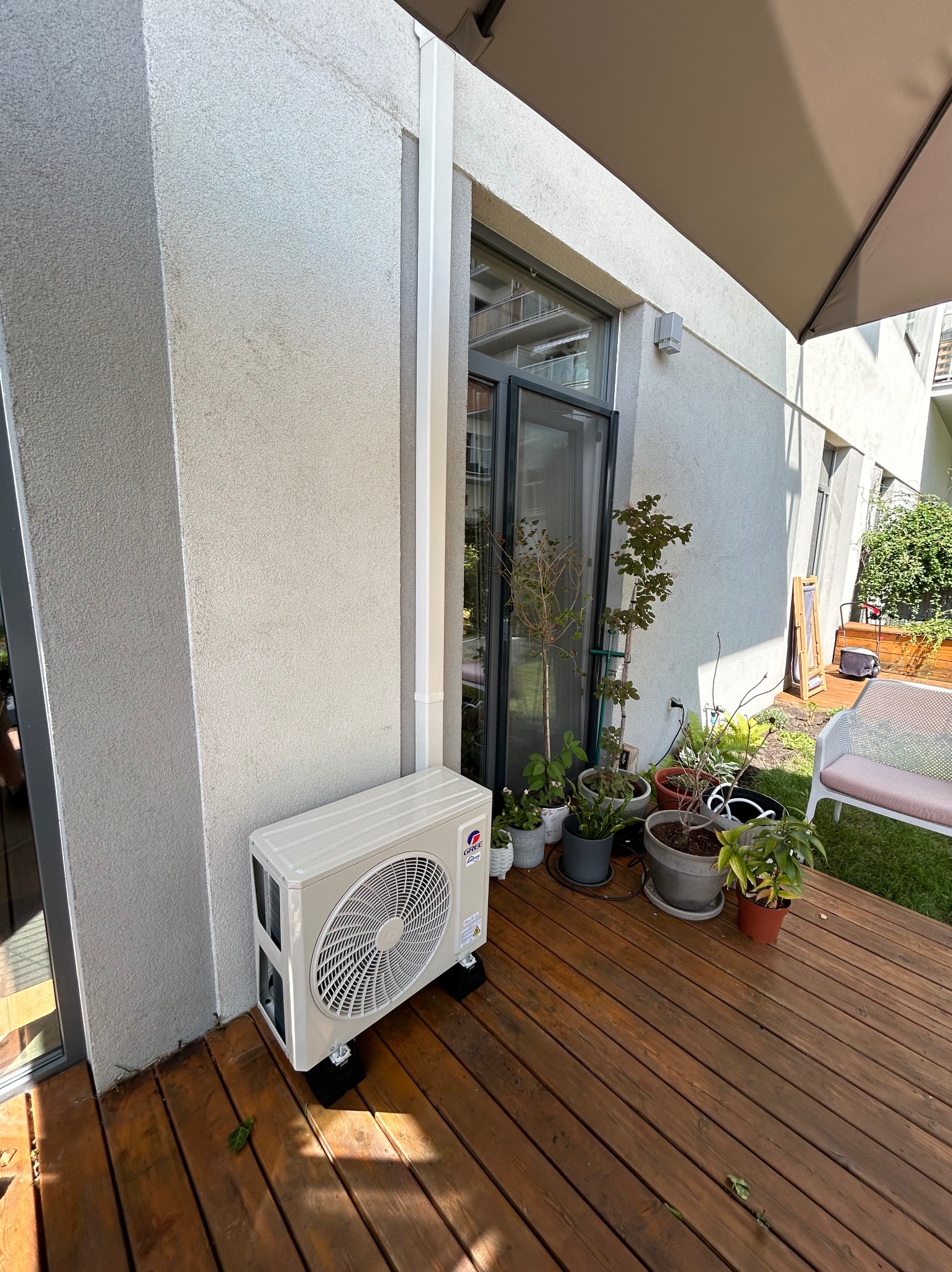 Biały klimatyzator zewnętrzny marki Gree zamontowany na drewnianym tarasie przy ścianie budynku, obok okna i donic z roślinami.