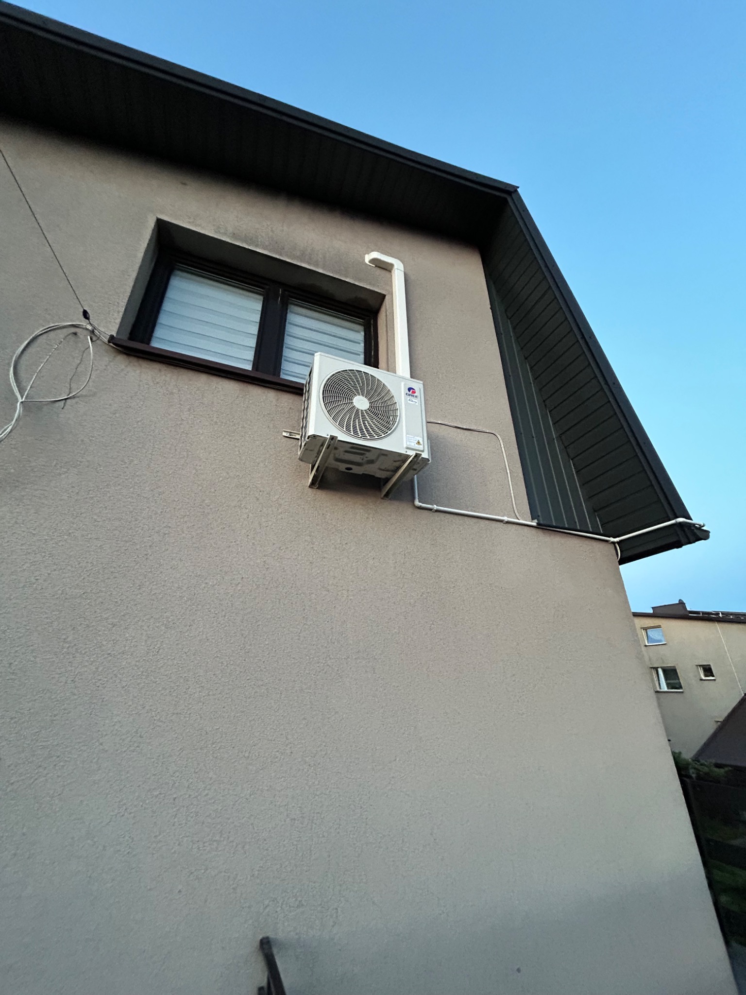 Zewnętrzna jednostka klimatyzacji GREE zamontowana na elewacji domu. Widoczne orurowanie i okno z żaluzjami. Kadr z dołu, błękitne niebo w tle.