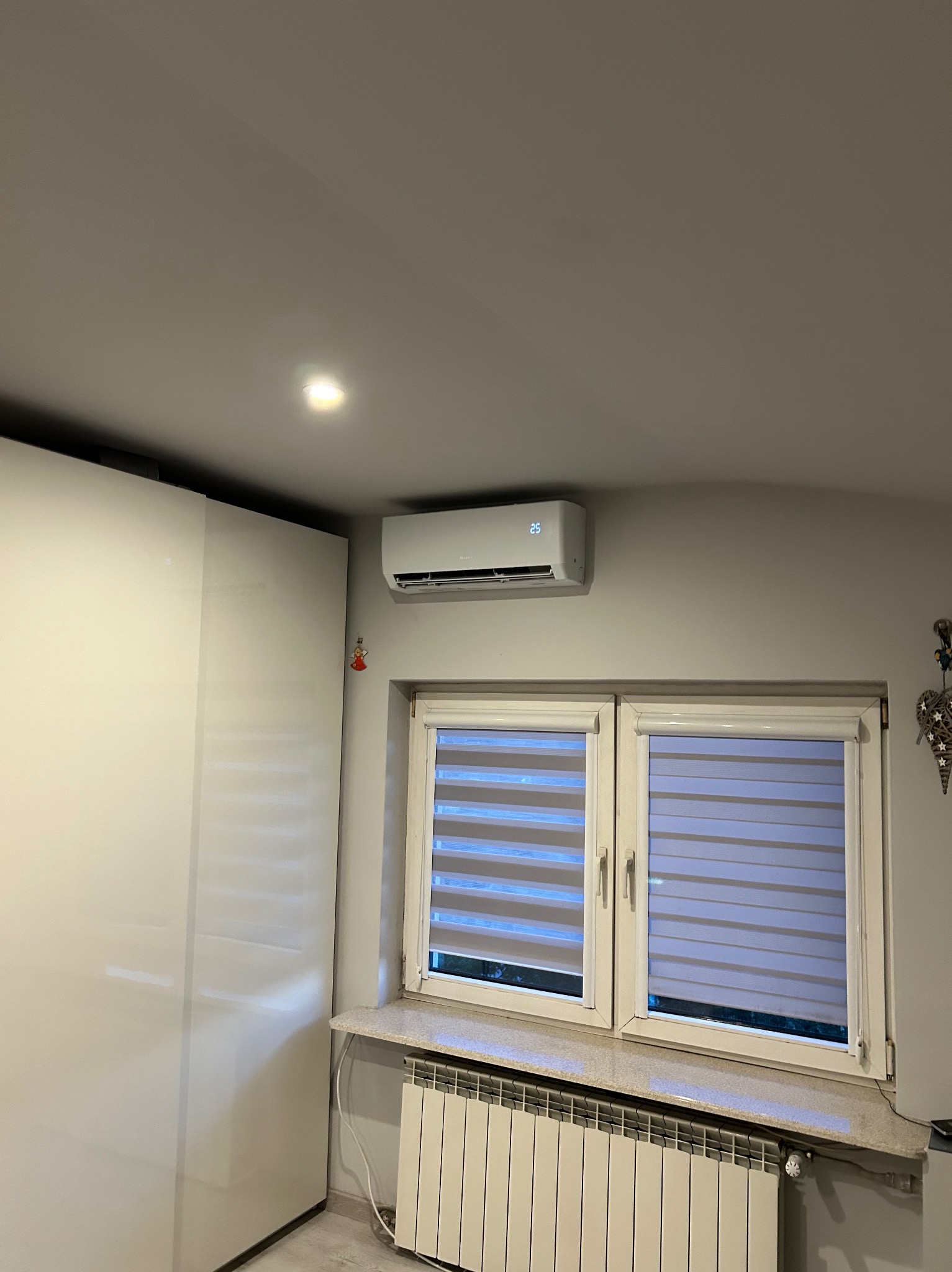 Biały klimatyzator ścienny z wyświetlaczem temperatury, zamontowany nad oknem z roletami w pokoju z grzejnikiem i białą szafą. Widok na fragment wnętrza.