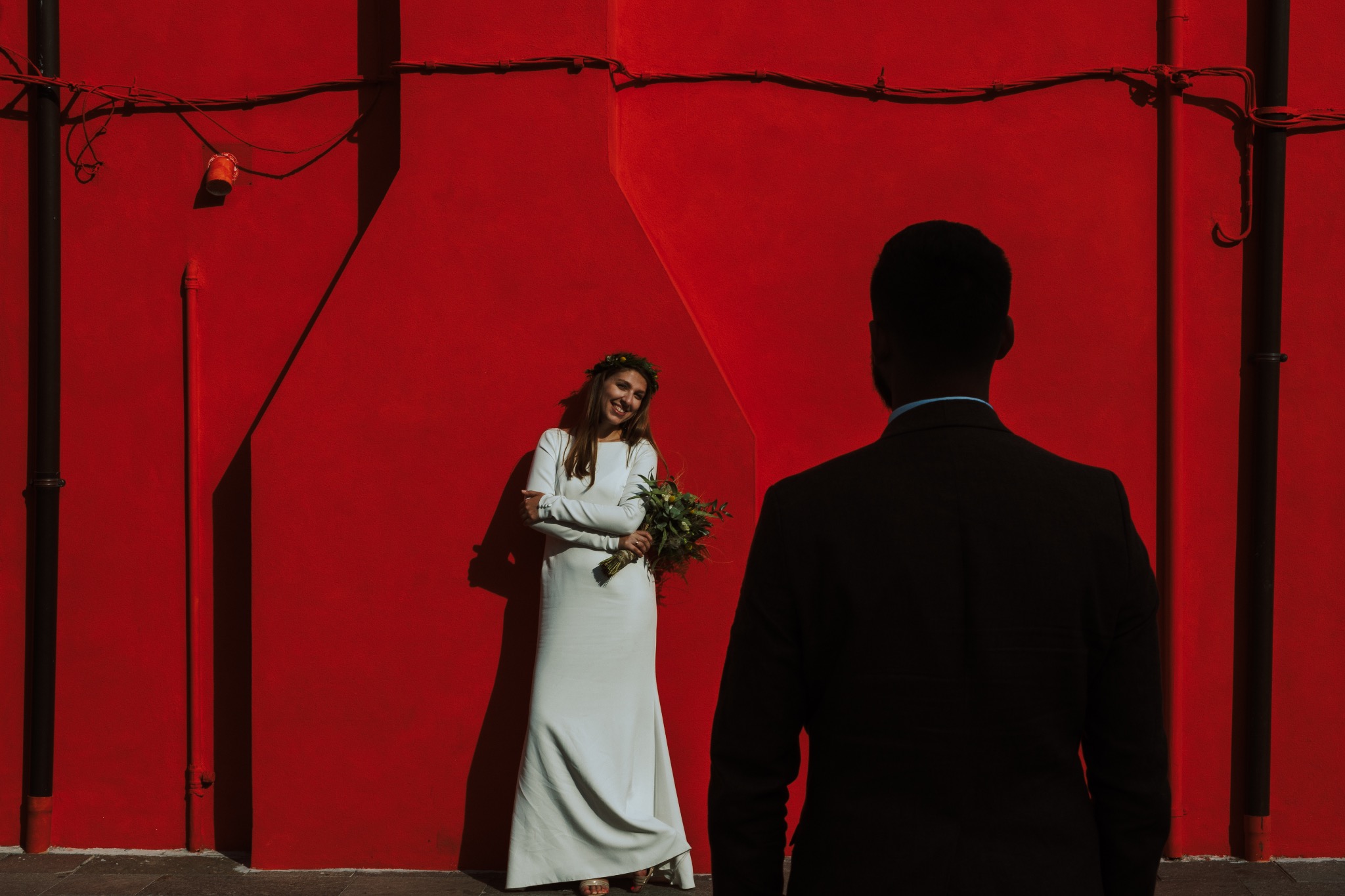 Panna młoda w białej sukni i wianku pozuje na tle czerwonej ściany, z boku widoczny mężczyzna w garniturze. Ujęcie z sesji ślubnej w intensywnych kolorach.