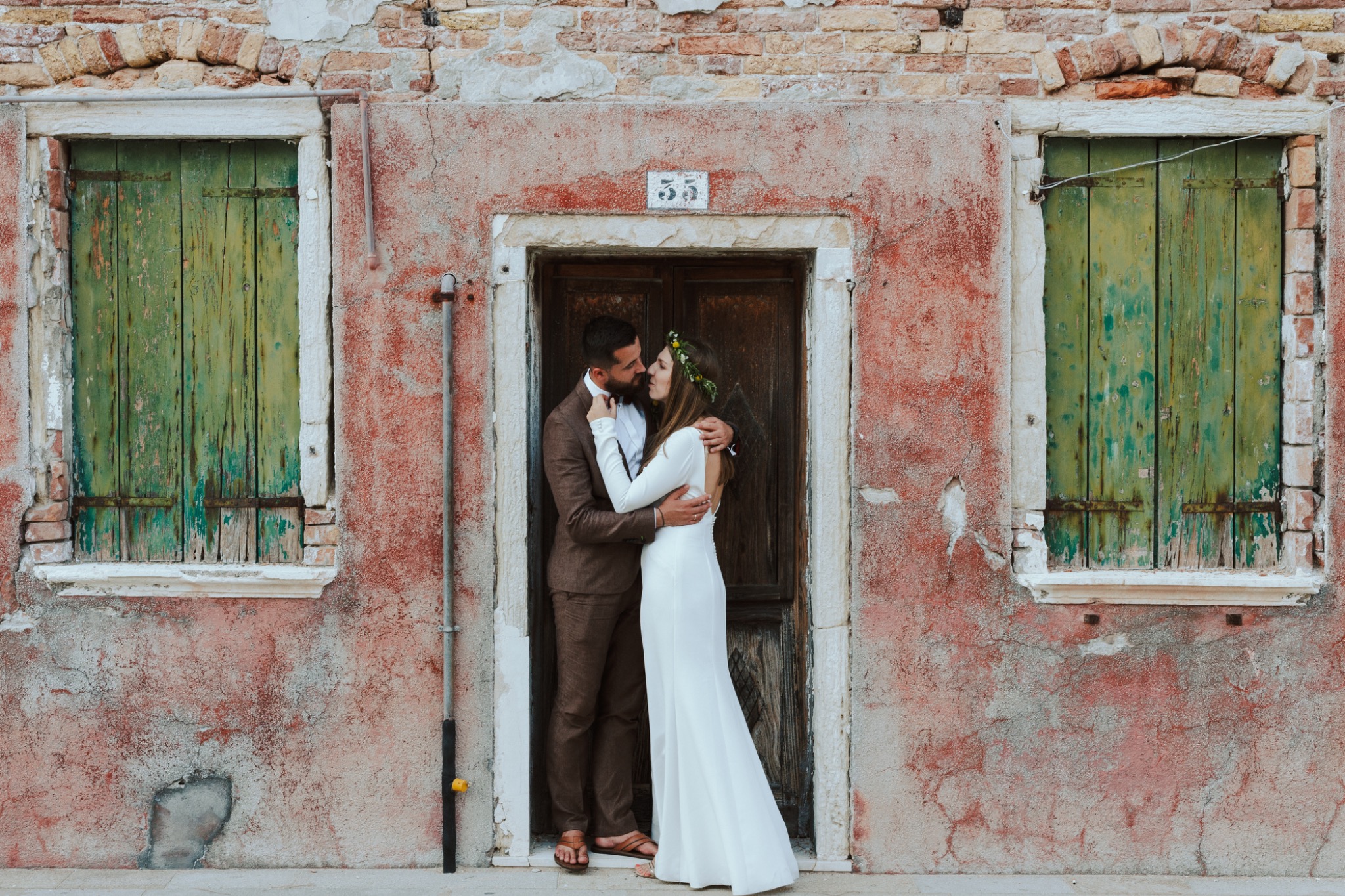 Para młoda w objęciach na tle starej, pomalowanej ściany z oknami z zielonymi okiennicami i numerem 55 nad drzwiami. Romantyczna sesja w rustykalnym stylu.