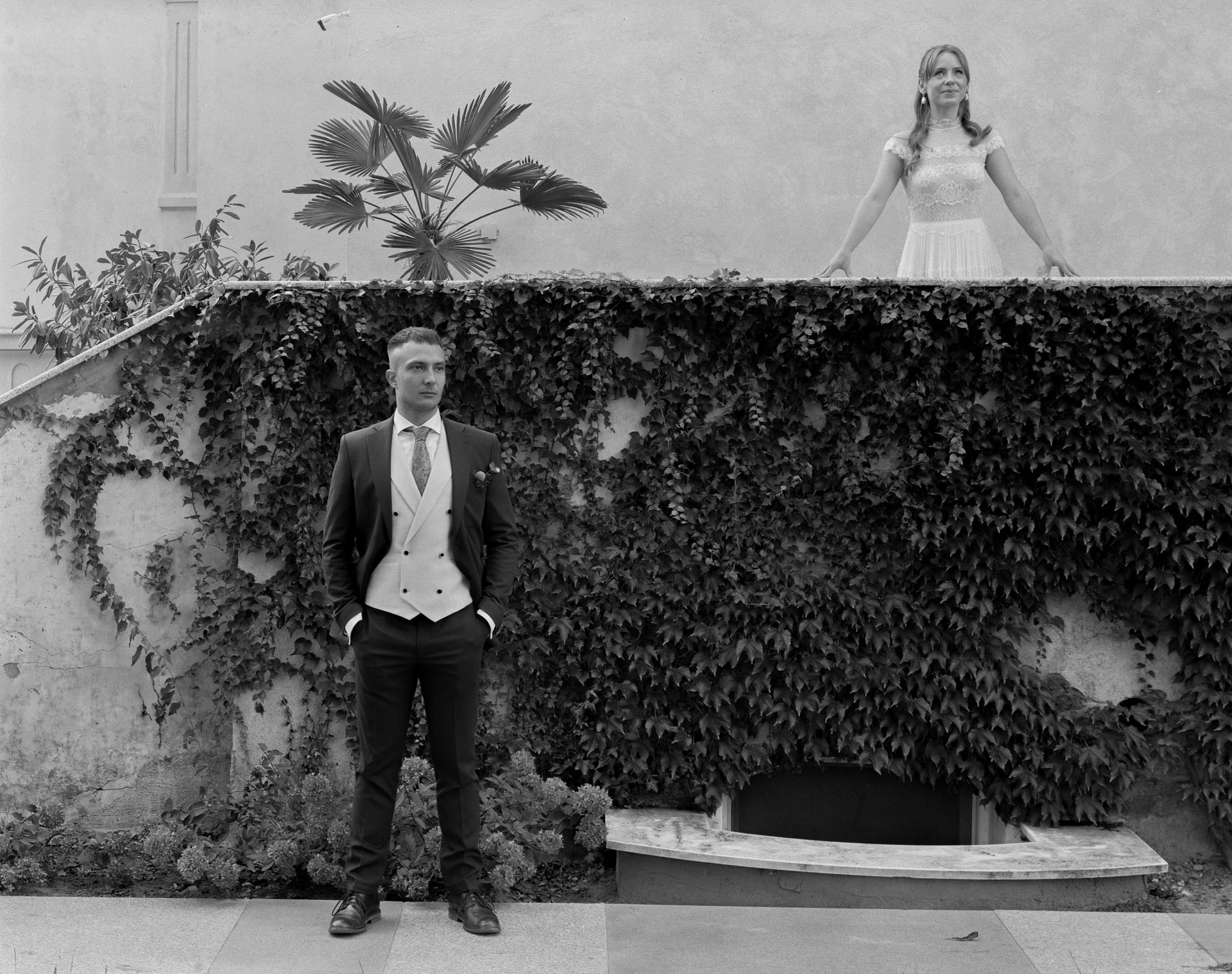 Czarno-białe zdjęcie pary młodej: pan młody w garniturze stoi na dole, a panna młoda opiera się o mur porośnięty bluszczem. Nietypowe ujęcie.