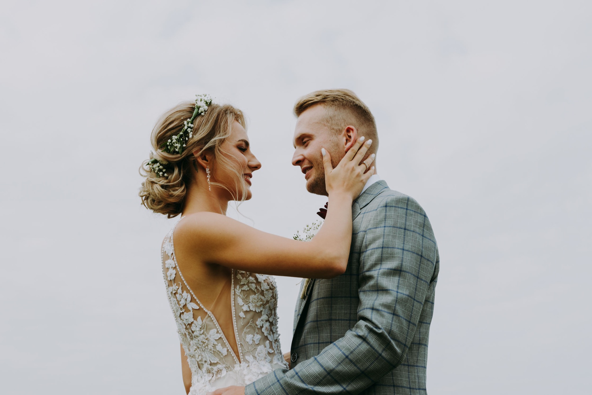 Młoda para wpatrzona w siebie podczas dnia ślubu. Panna młoda z wiankiem i koronkową suknią, pan młody w kraciastej marynarce. Delikatne, naturalne światło.