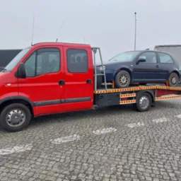 Czerwona laweta Renault z załadowanym, ciemnym hatchbackiem na platformie, na parkingu z kostki brukowej w pochmurny dzień. Widoczne elementy oświetlenia i inne pojazdy w tle.