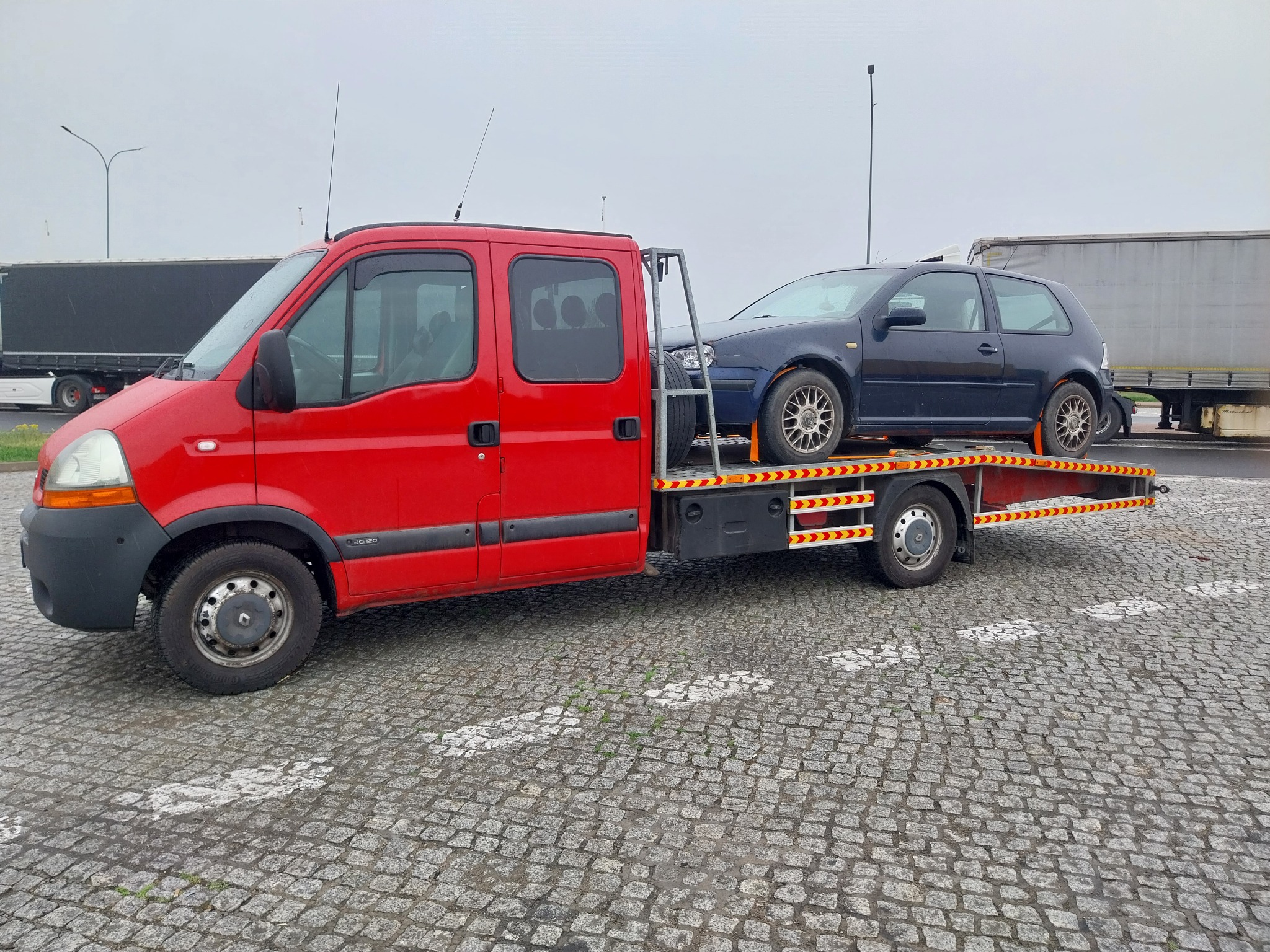 Czerwona laweta Renault z załadowanym, ciemnym hatchbackiem na platformie, na parkingu z kostki brukowej w pochmurny dzień. Widoczne elementy oświetlenia i inne pojazdy w tle.