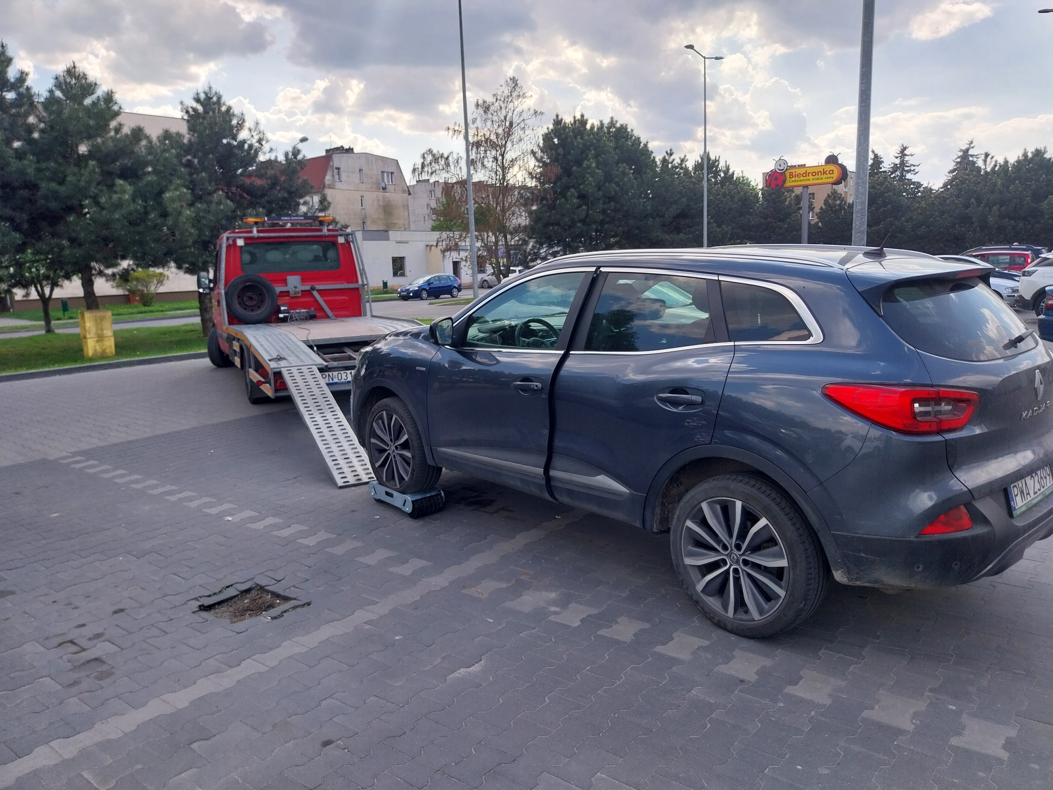 Ciemny SUV na wózku lawety, otwarte drzwi, w tle czerwona laweta z rampą, parking z kostki, w tle budynek i reklama Biedronki.