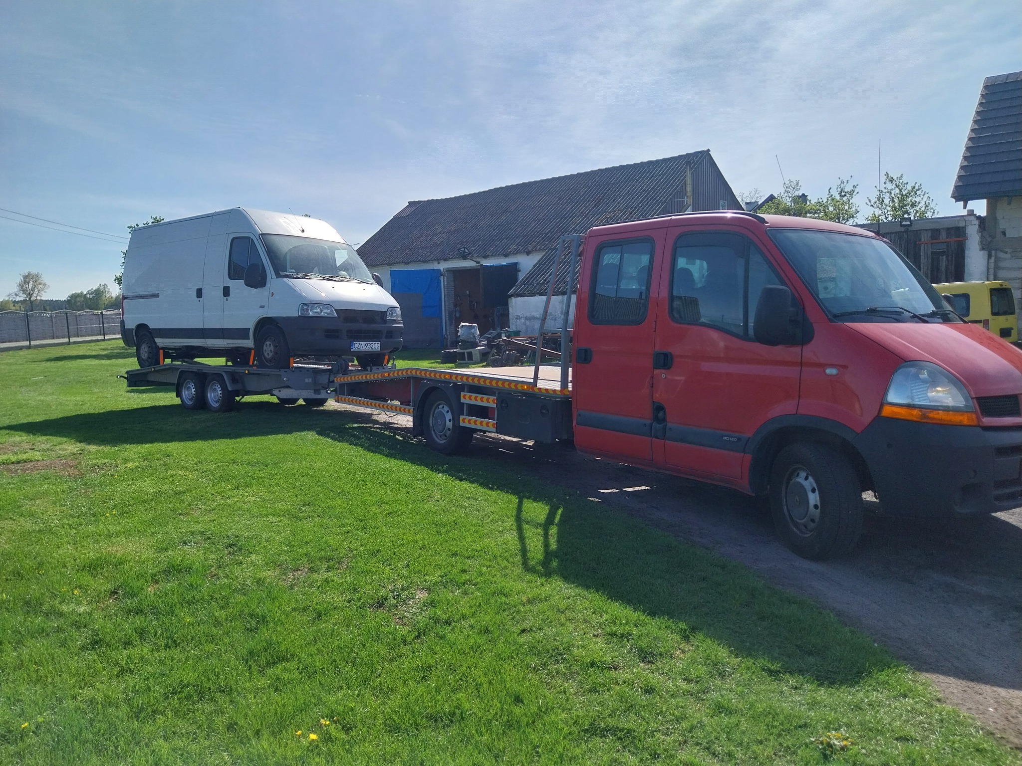 Czerwona platforma transportowa z białym vanem na lawecie, zaparkowana na trawniku przed budynkiem gospodarczym w słoneczny dzień.