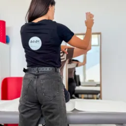 Osoba w czarnej koszulce z logo RehaBil wykonuje ćwiczenia z pacjentem na leżance w jasnym gabinecie rehabilitacyjnym. Widok z tyłu.