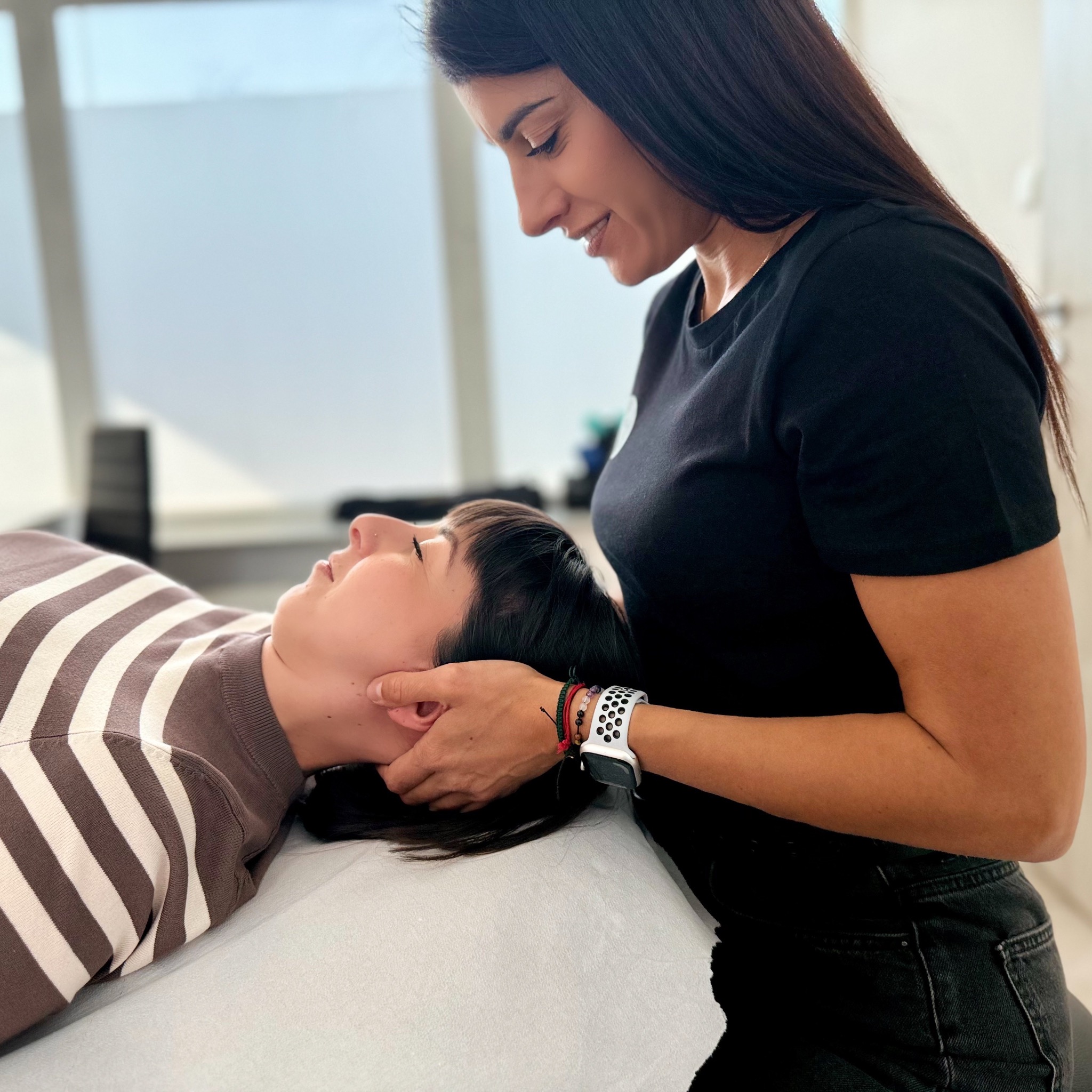 Kobieta poddawana terapii manualnej szyi przez terapeutkę w czarnym T-shircie, leżąca na jasnym podłożu, z widocznym zegarkiem sportowym.