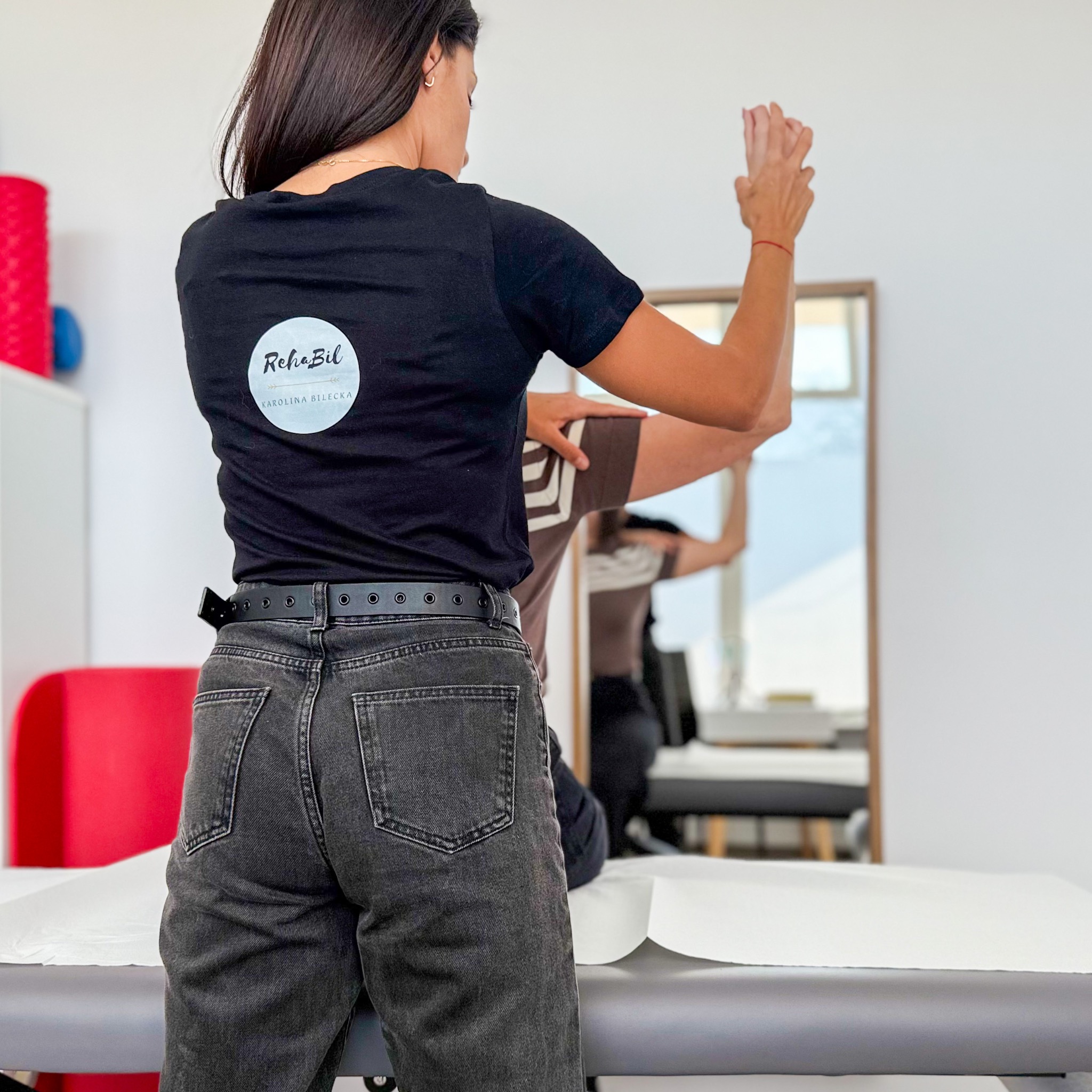 Osoba w czarnej koszulce z logo RehaBil wykonuje ćwiczenia z pacjentem na leżance w jasnym gabinecie rehabilitacyjnym. Widok z tyłu.