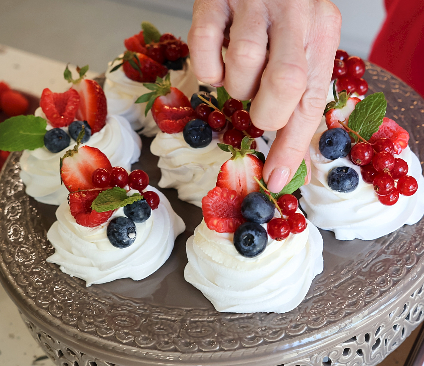 Eleganckie mini bezy Pavlova z świeżymi owocami (truskawki, borówki, porzeczki) i miętą, układane na ozdobnej paterze przez kobiecą dłoń.