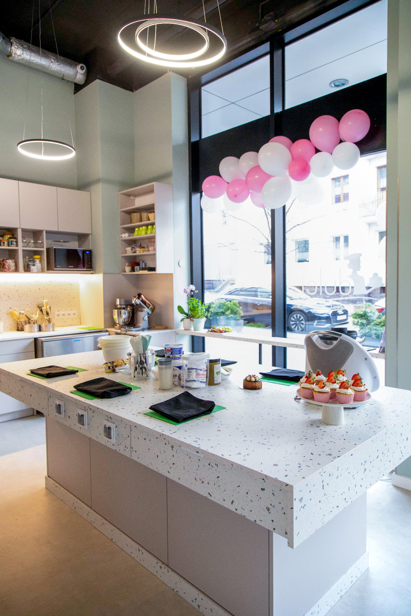 Jasna kuchnia cukiernicza z wyspą, na której leżą składniki, babeczki ze świeżymi truskawkami i dekoracja z różowych balonów przy oknie.