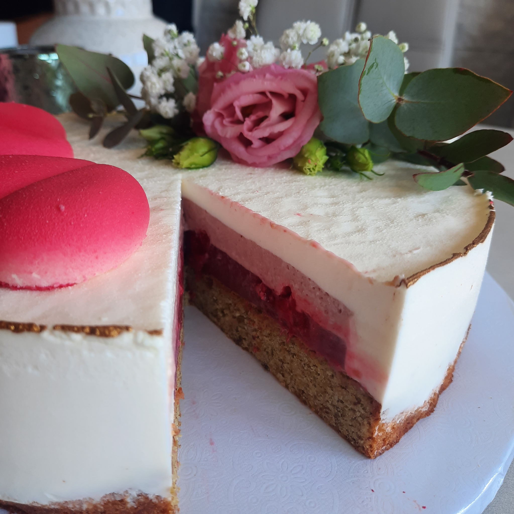 Kremowy tort z różowymi dekoracjami kwiatowymi i makaronikami, z wyciętym kawałkiem, ukazującym warstwy ciasta i kremu.