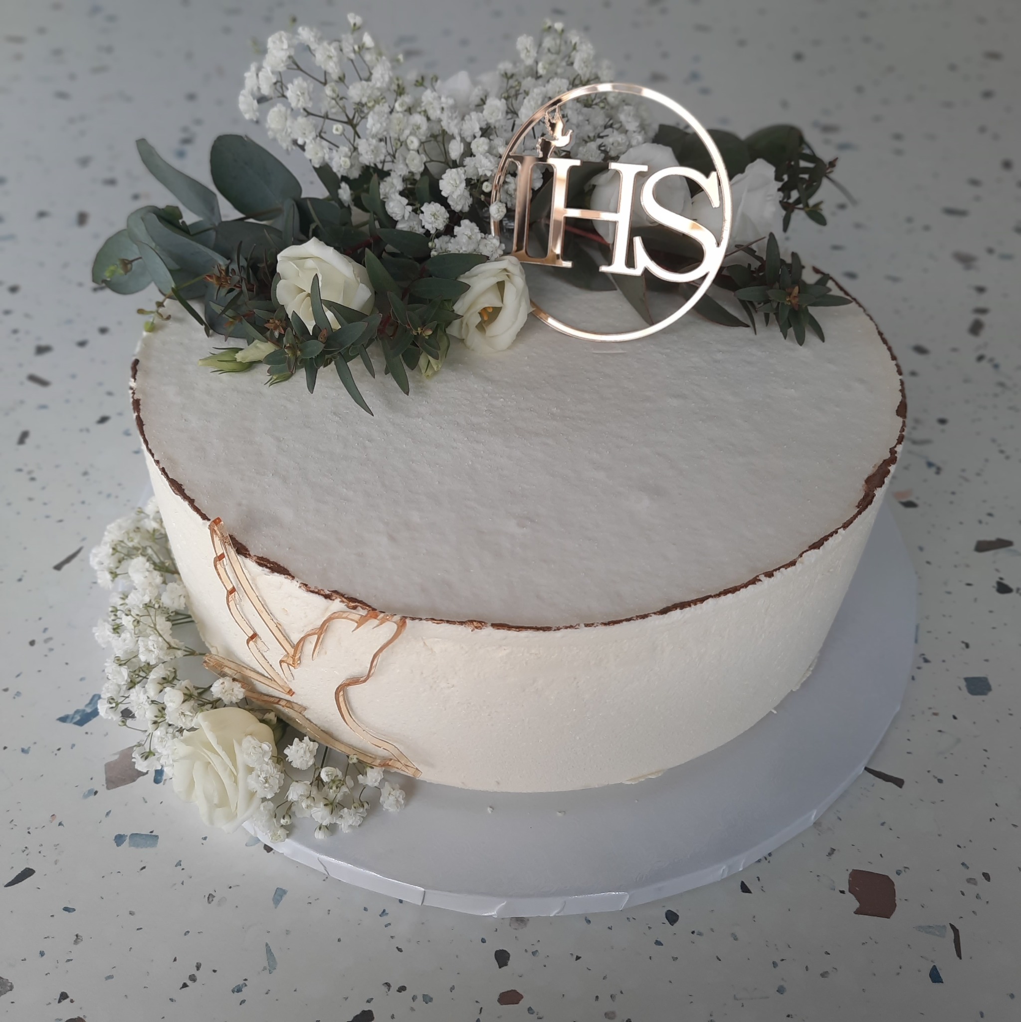 Elegancki tort komunijny z białym kremem, złotym topperem IHS, delikatnymi kwiatami i eukaliptusem na tle lastryko.