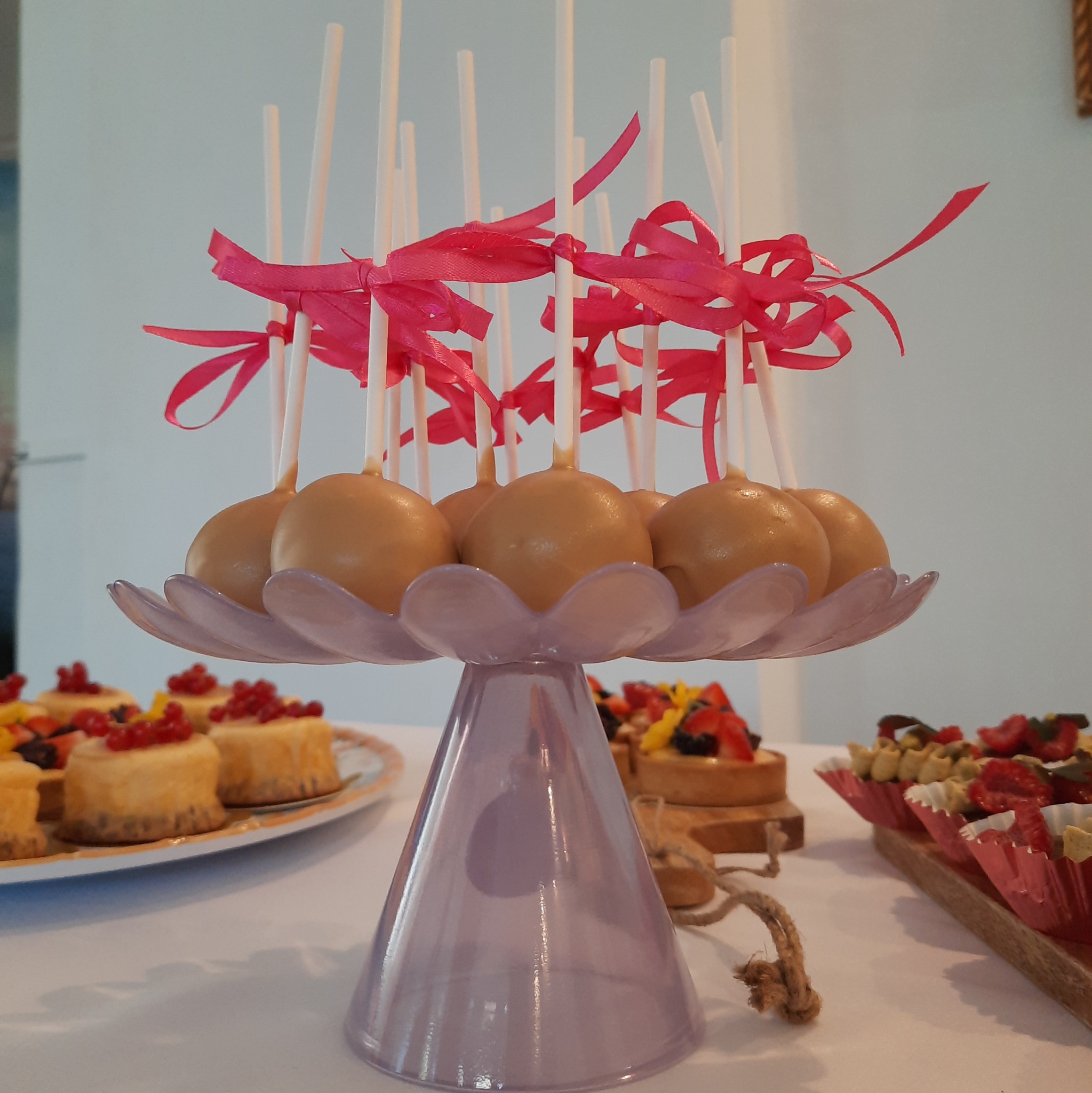 Karmelowe cake pops z różowymi wstążkami na fioletowej paterze, obok mini serniczki i tartaletki z owocami. Słodki stół na przyjęcie.