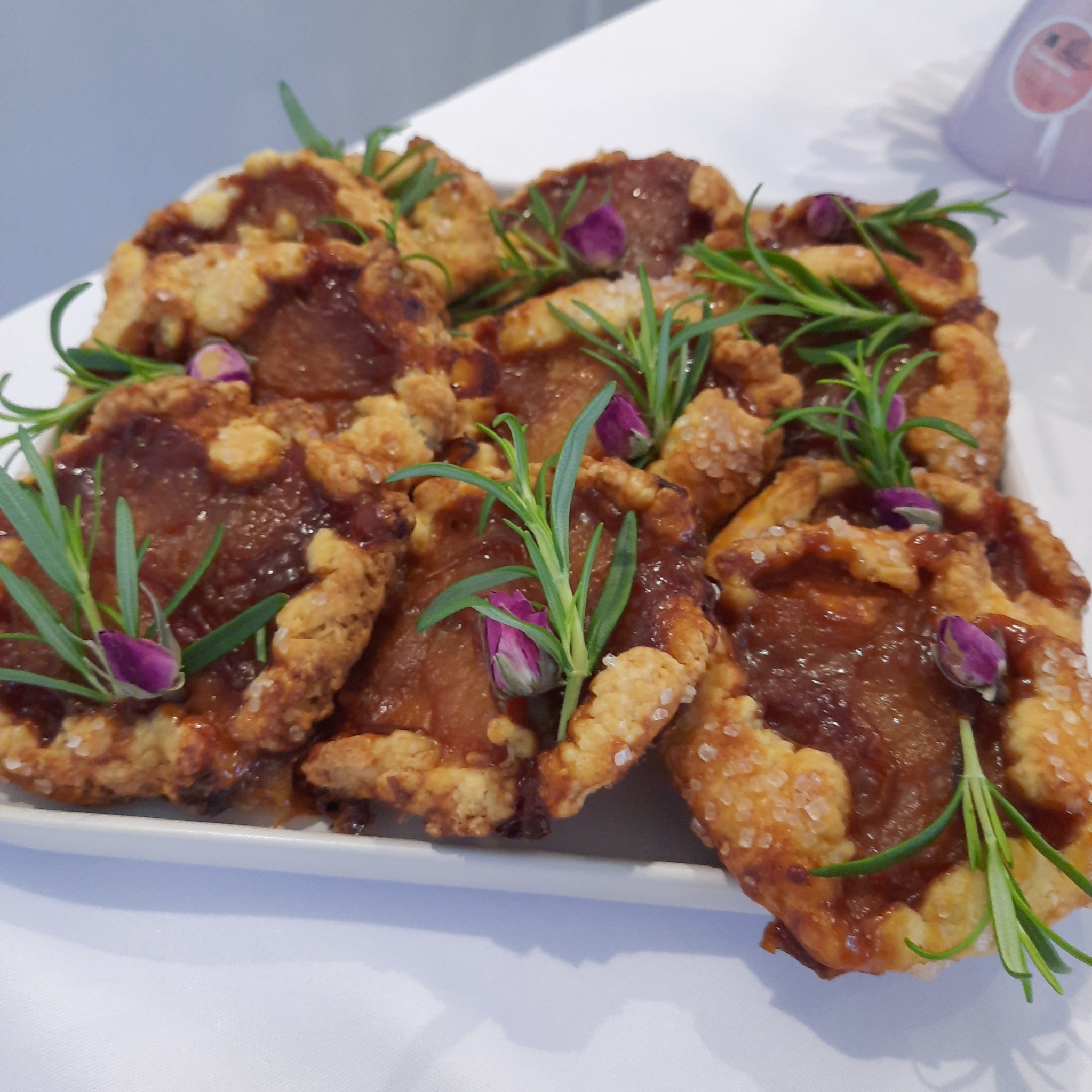 Rustykalne tartaletki z jabłkami, udekorowane gałązkami rozmarynu i pąkami róż, ułożone na białym talerzu. Widoczne kryształki cukru na cieście.