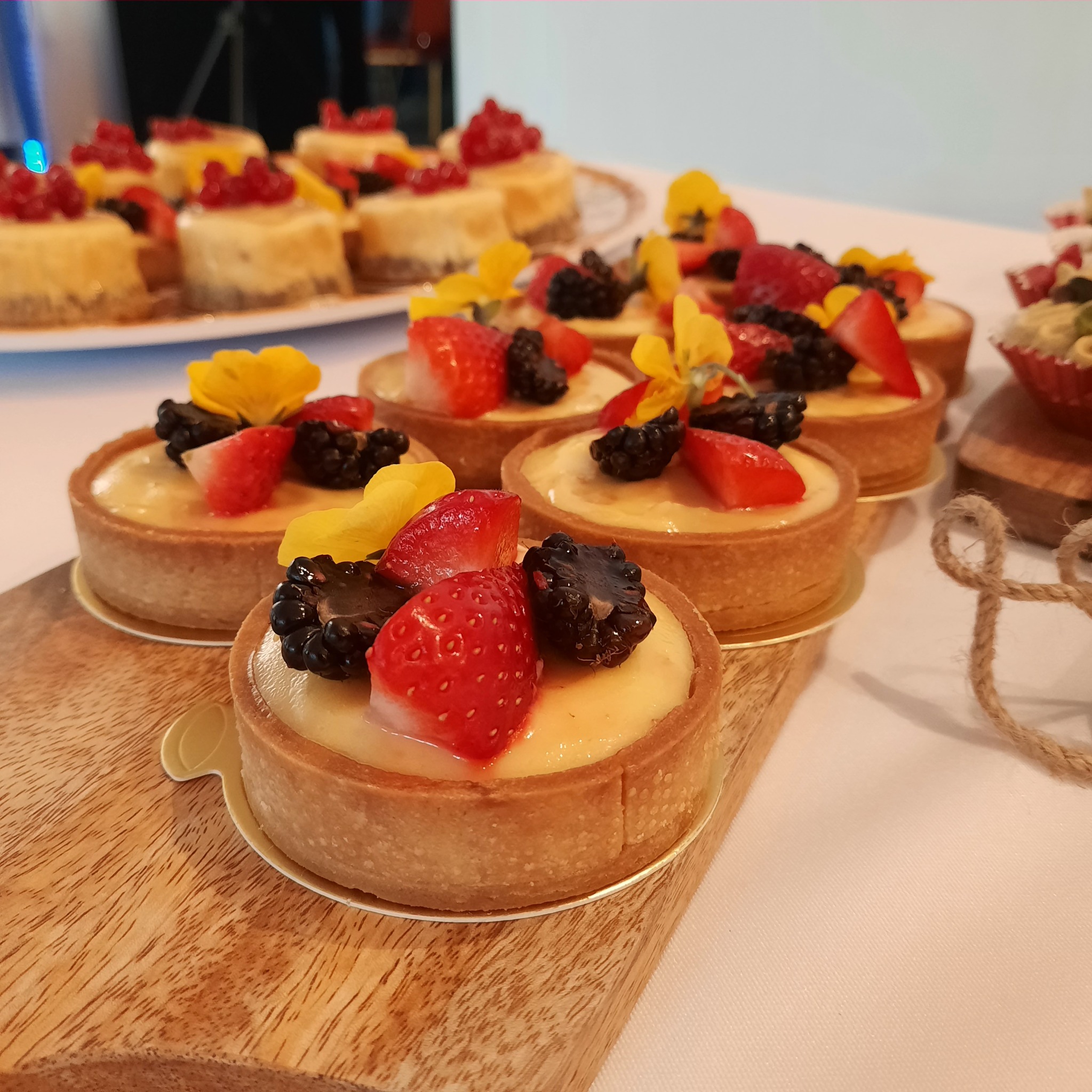 Mini tartaletki z kremem, truskawkami, jeżynami i jadalnymi kwiatami na drewnianej desce. W tle inne desery.