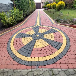 Piotr Wątroba - Kolorowa kostka brukowa ułożona w ozdobny wzór, prowadząca wzdłuż zadbanego ogrodu i betonowego ogrodzenia w kierunku bramy garażowej.
