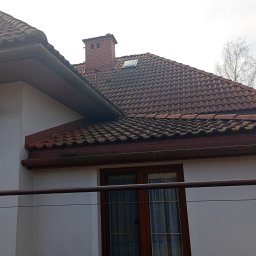 Mycie ciśnieniowe - Ceglany komin i dach z dachówki ceramicznej na tle jasnej elewacji domu. Widoczne rynny i okno. Ujęcie z dołu, podkreśla strukturę i detale pokrycia dachowego.