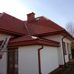 Dom z nowym, czerwonym dachem z dachówki ceramicznej i rynnami w tym samym kolorze. Przystawiona drabina sugeruje prace dekarskie.