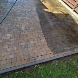 Częściowo umyta kostka brukowa w odcieniach brązu, widoczne różnice w czystości, zarys węża ciśnieniowego i cień drabiny, trawa wzdłuż krawędzi.