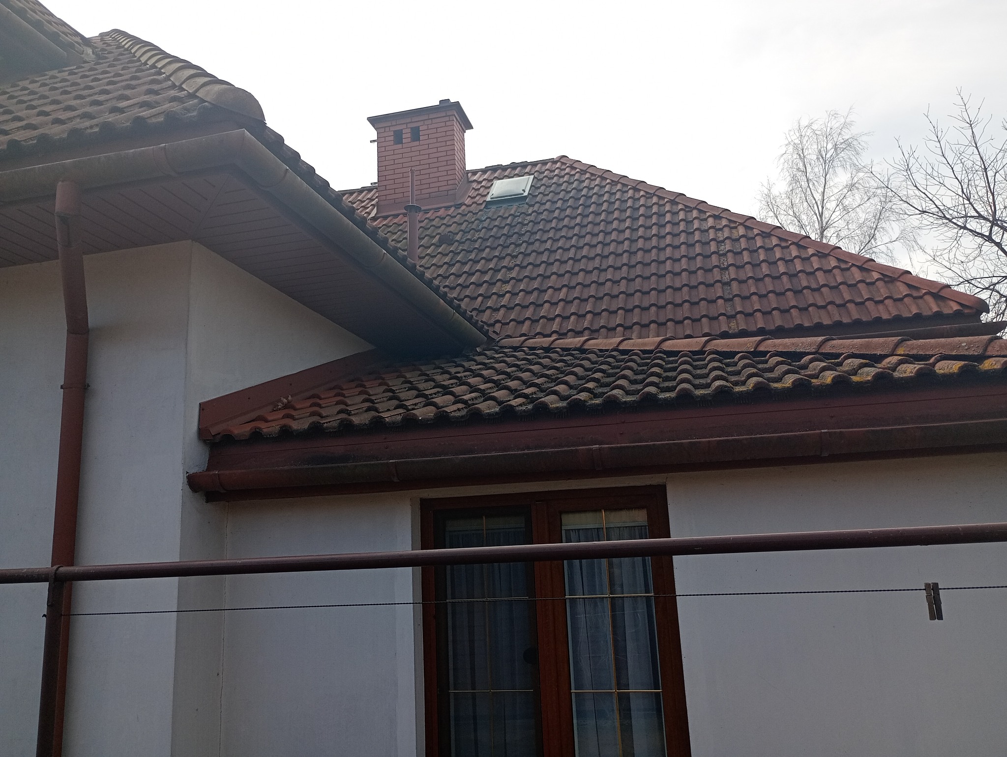 Ceglany komin i dach z dachówki ceramicznej na tle jasnej elewacji domu. Widoczne rynny i okno. Ujęcie z dołu, podkreśla strukturę i detale pokrycia dachowego.