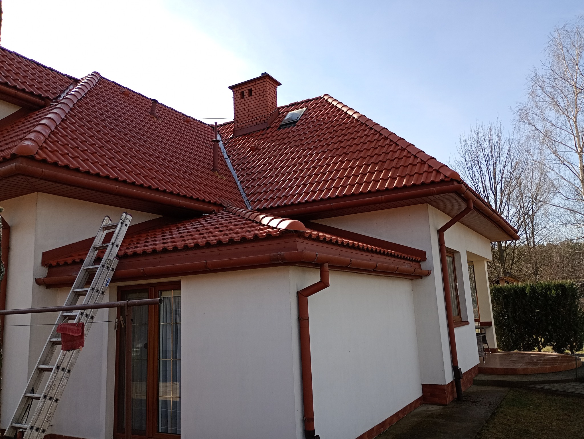 Dom z nowym, czerwonym dachem z dachówki ceramicznej i rynnami w tym samym kolorze. Przystawiona drabina sugeruje prace dekarskie.