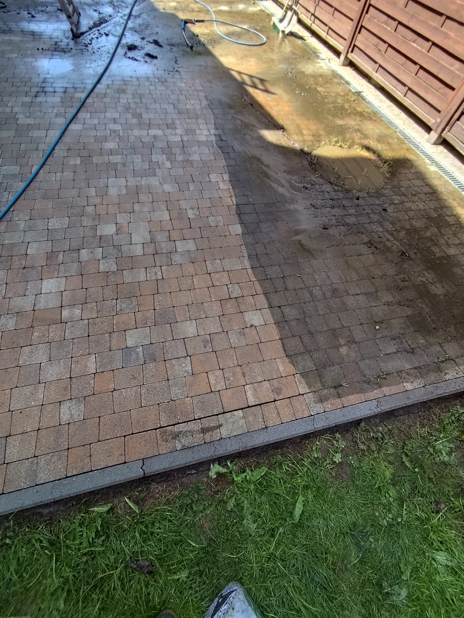 Częściowo umyta kostka brukowa w odcieniach brązu, widoczne różnice w czystości, zarys węża ciśnieniowego i cień drabiny, trawa wzdłuż krawędzi.