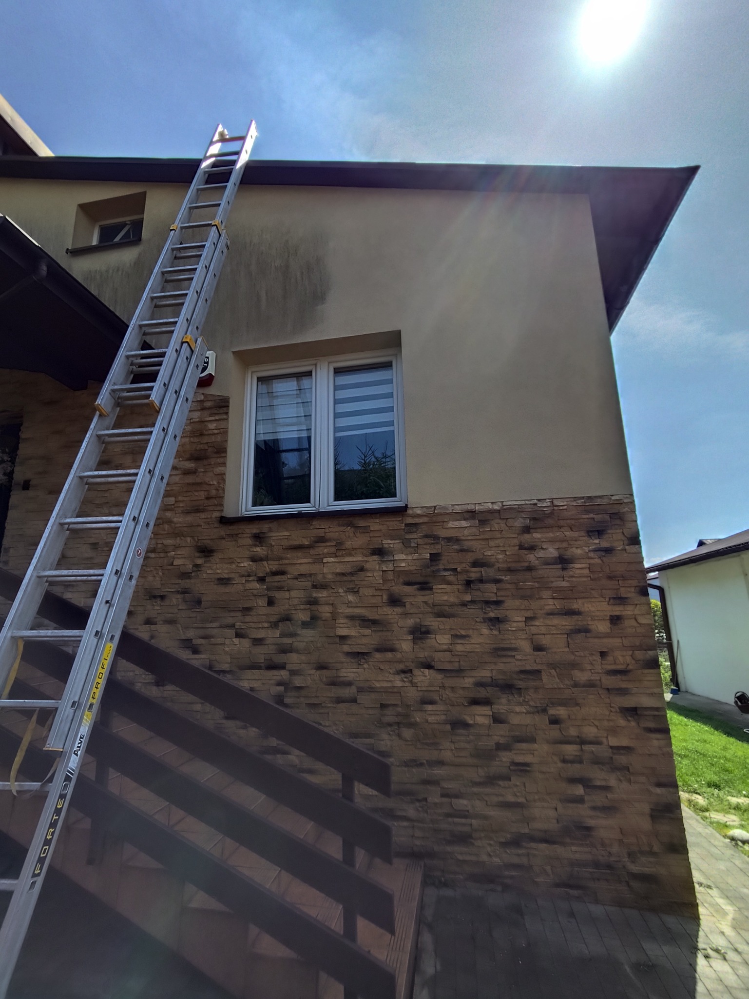 Dwupiętrowy dom z elewacją częściowo z kamienia, z widocznymi zabrudzeniami na górnej, jasnej części ściany. Przystawiona drabina sugeruje prace na wysokości.