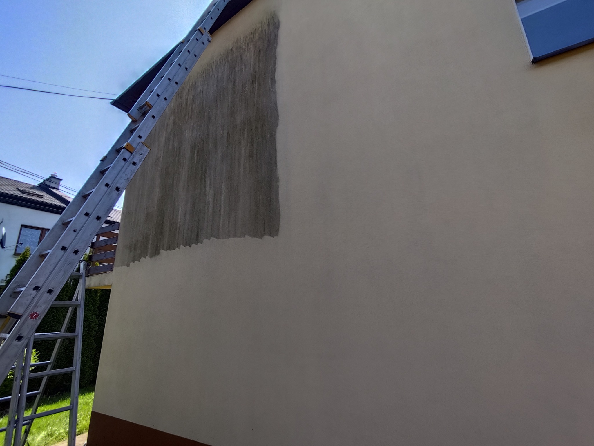 Fragment elewacji budynku w trakcie mycia, widoczna drabina oparta o ścianę. Część ściany umyta, reszta jeszcze zabrudzona. Kontrast czystej i brudnej powierzchni.