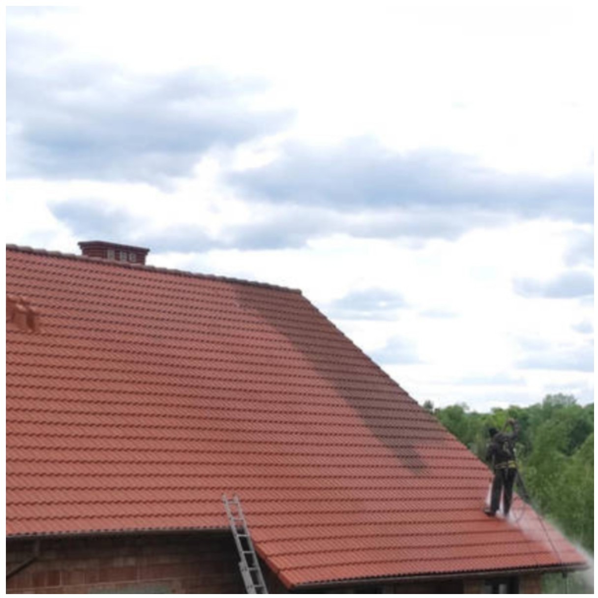 Mycie dachu z czerwonej dachówki. Widoczna różnica czystości, drabina i pracownik w uprzęży, spryskujący dach wodą pod ciśnieniem. Częściowo zachmurzone niebo w tle.