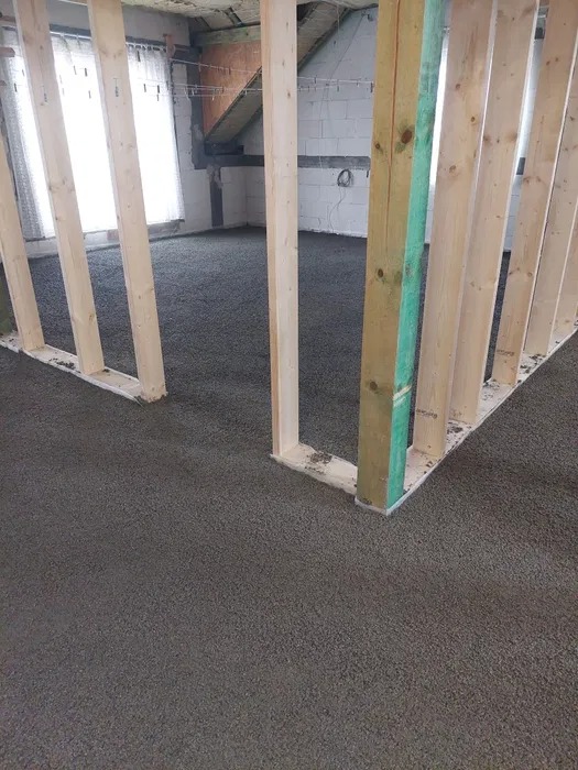 Wylany jastrych cementowy wewnątrz budynku, widoczne drewniane elementy konstrukcyjne. Szary jastrych tworzy równą powierzchnię. W tle okno i ściana z białego pustaka.