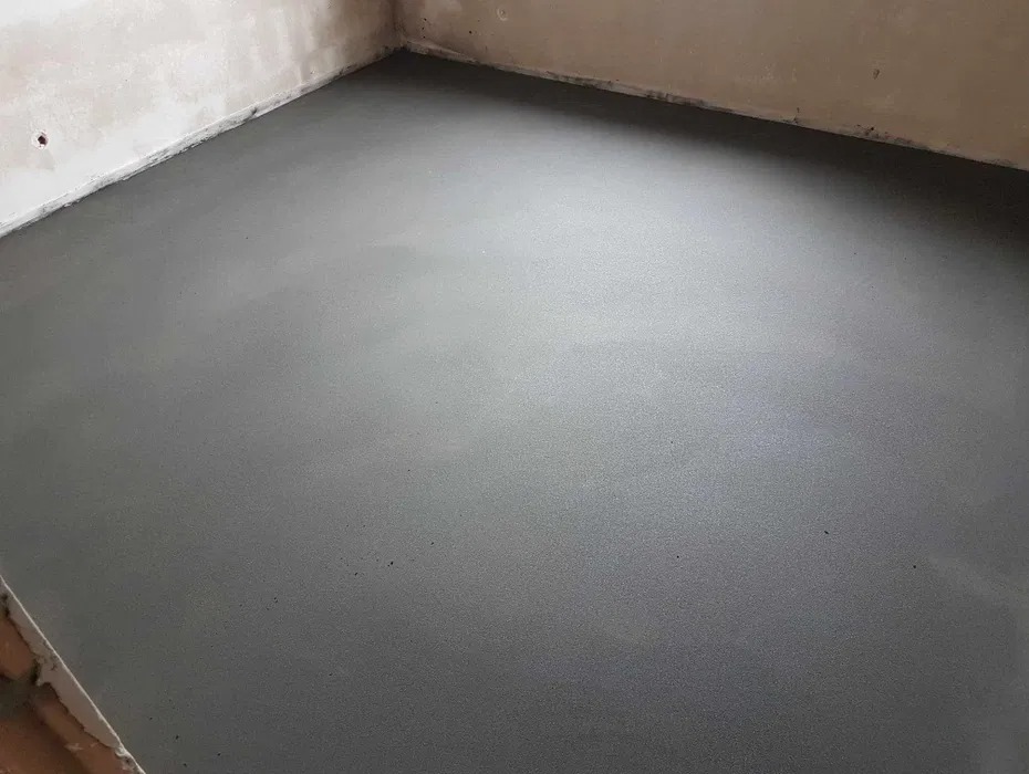 Gładka, szara posadzka jastrychowa w surowym wnętrzu. Widoczne niedoskonałości ścian i narożników. Minimalistyczny kadr, neutralne oświetlenie.