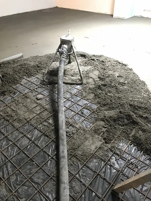 Wylewka jastrychowa: szara masa na zbrojeniu, wąż pompy w centrum kadru. Częściowo zalana podłoga w tle. Wnętrze w trakcie prac wykończeniowych.