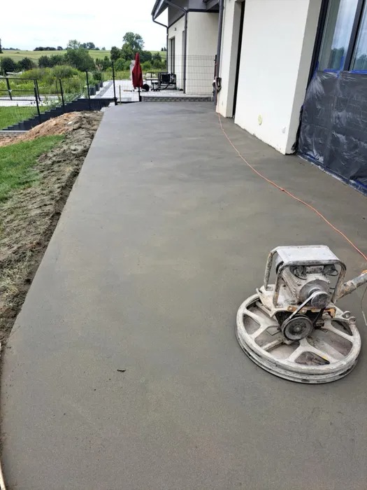 Wygładzona, szara wylewka betonowa tarasu przy domu jednorodzinnym, obok stoi szlifierka do betonu. Widok na ogród w tle.